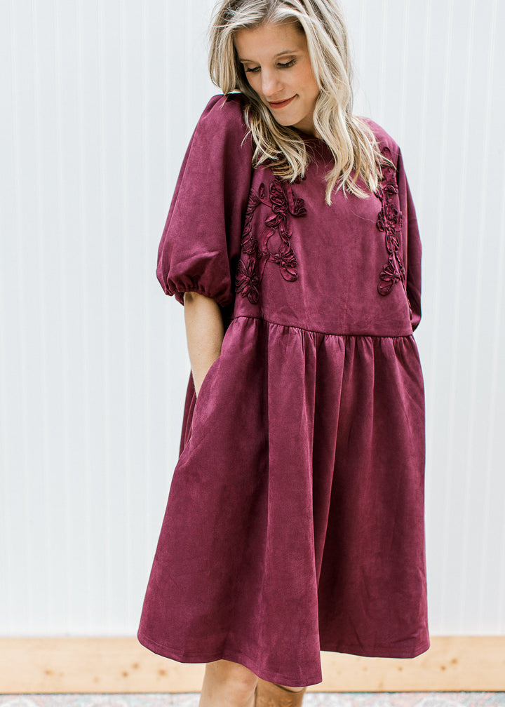 Model wearing a suede burgundy dress with floral detail, pockets and elbow length bubble sleeves.
