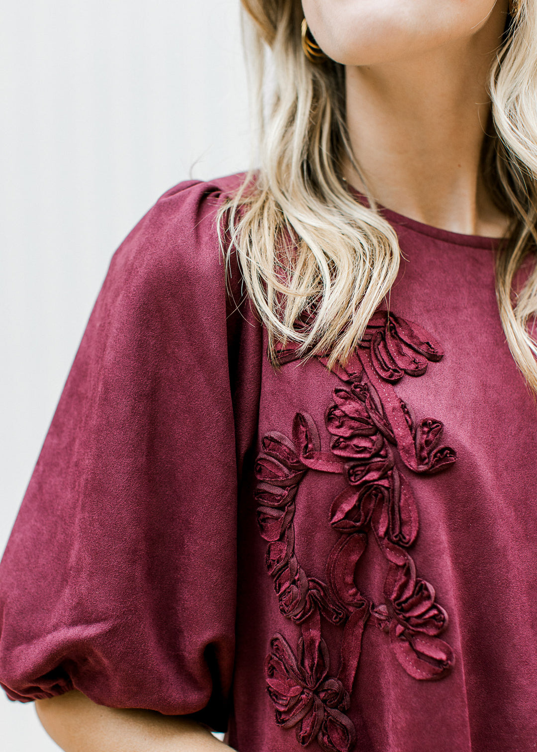 Close up view of floral detail, round neck and pleated shoulder on a suede burgundy dress. 
