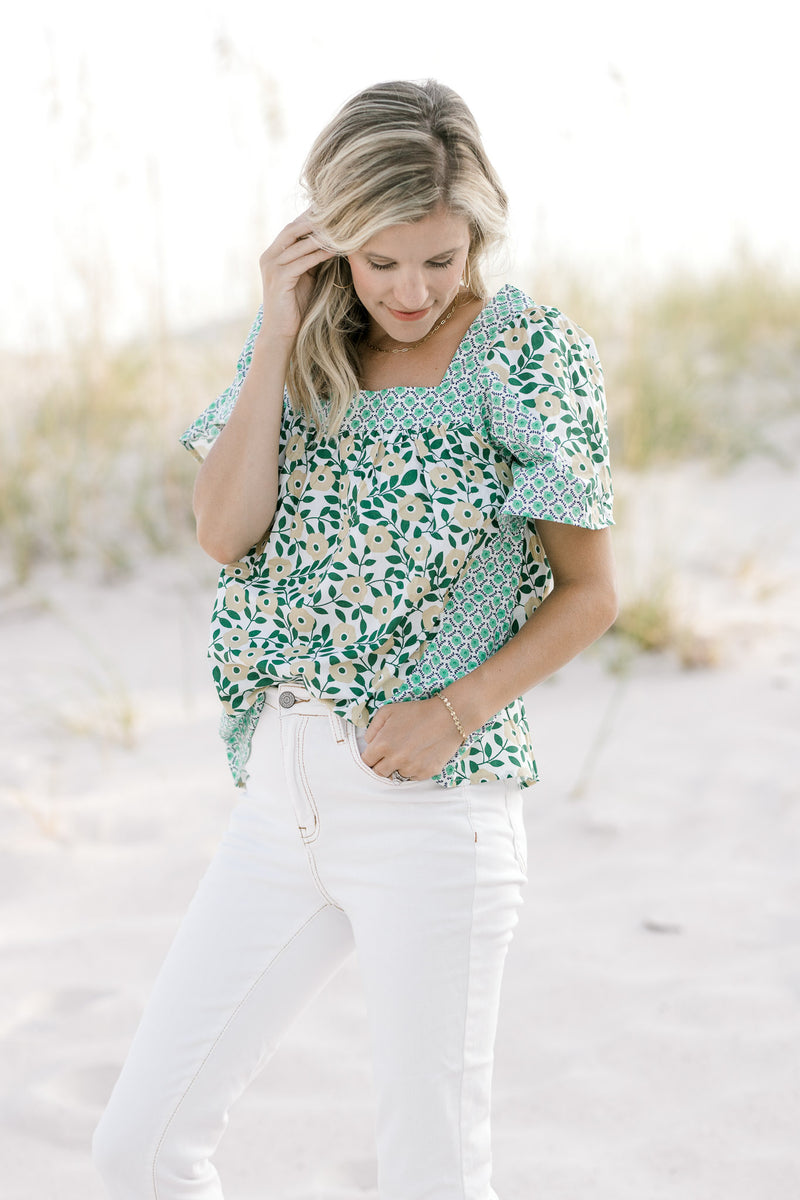 Model wearing a mixed green floral pattern top with a square neckline tucked into white pants.