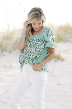 Model wearing a mixed green floral pattern top with a square neckline tucked into white pants.