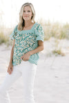 Model wearing white jeans with a mixed green floral top with a square neck and short sleeves. 