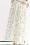 Model wearing a white maxi skirt with a cream floral pattern that is pleated with an elastic waist. 