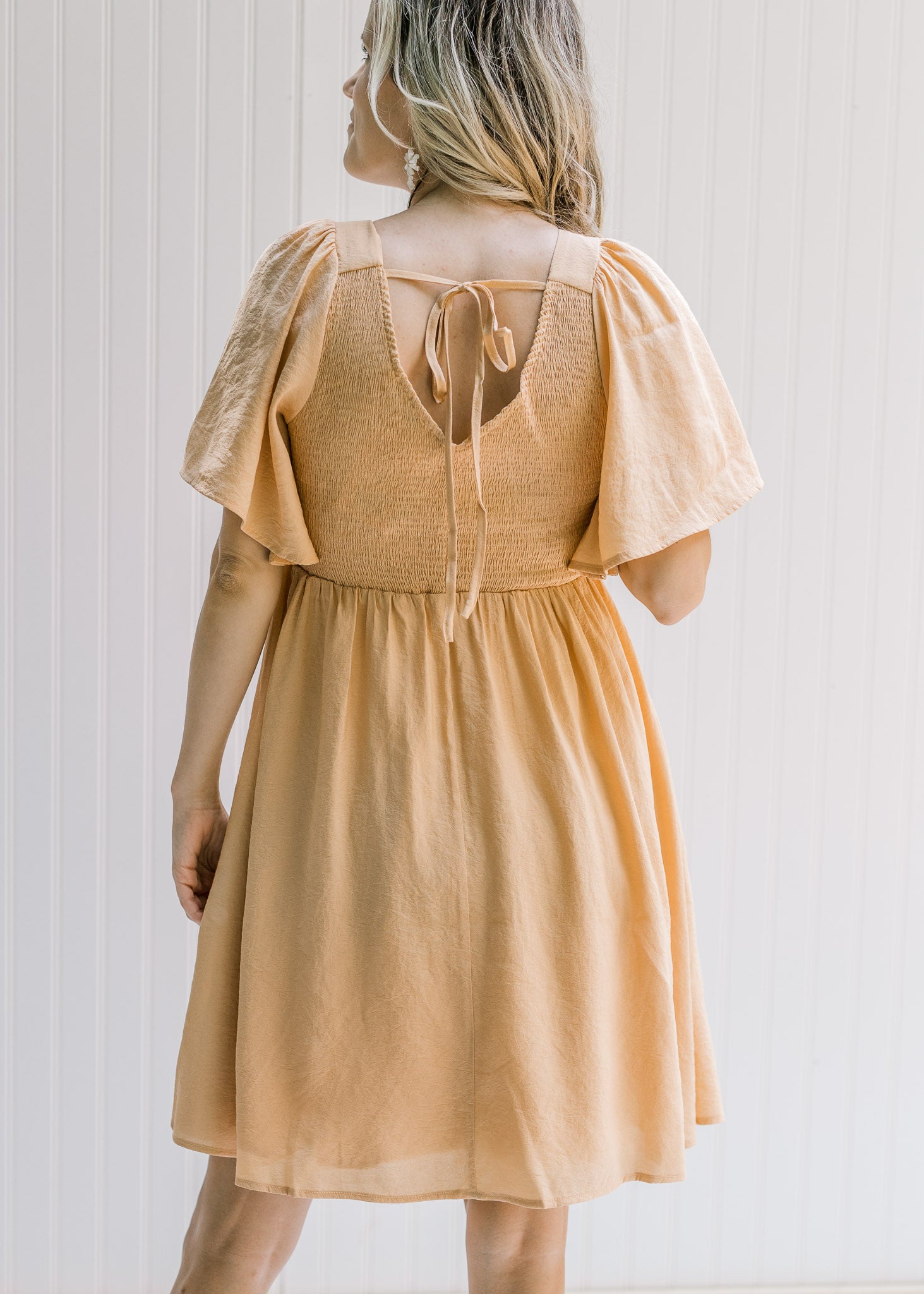 Back view of a model wearing a salmon dress with a smocked bodice, short sleeves and a tie closure. 