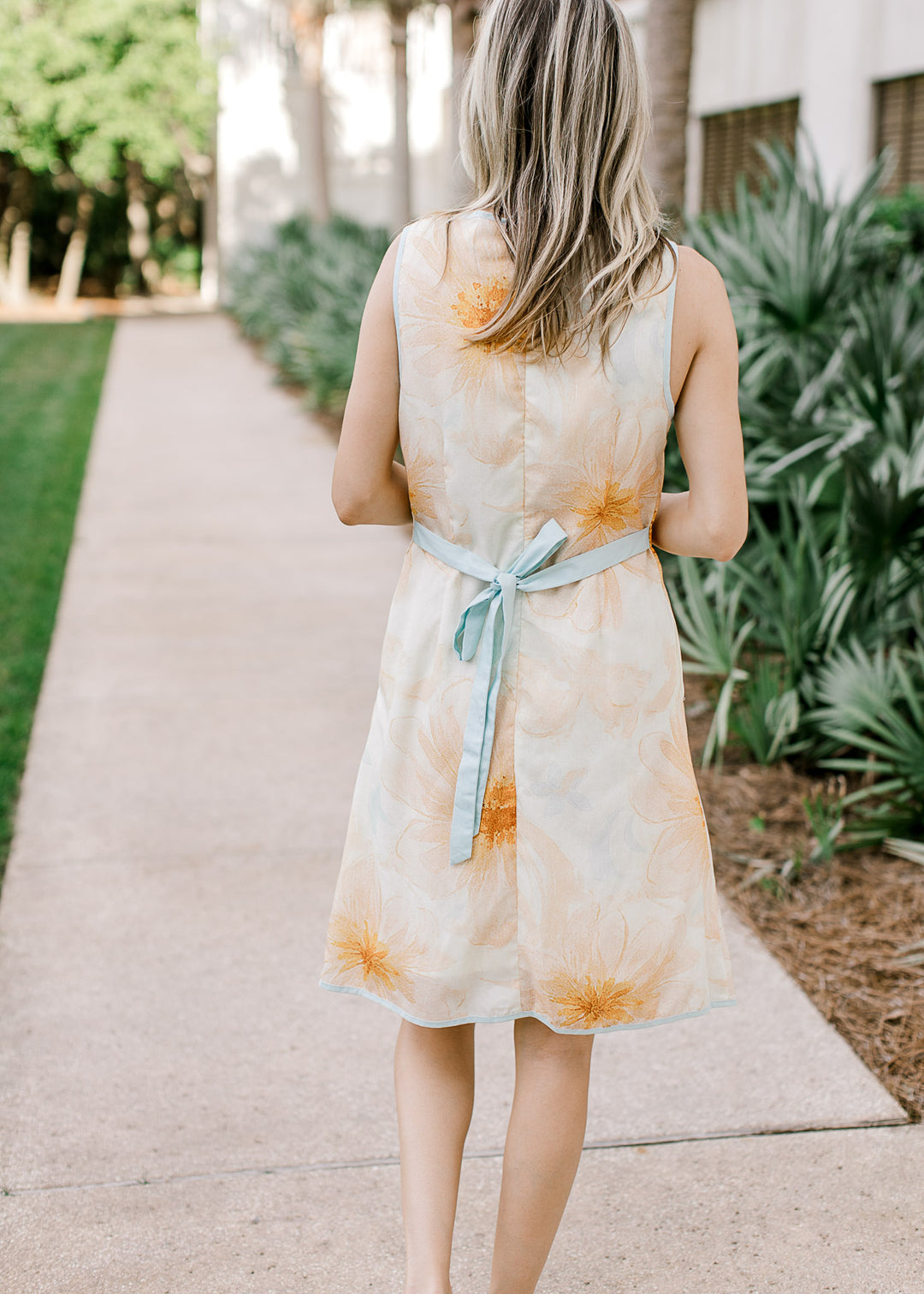 Back view of Model wearing a cream dress with orange and baby blue floral and baby blue tie.