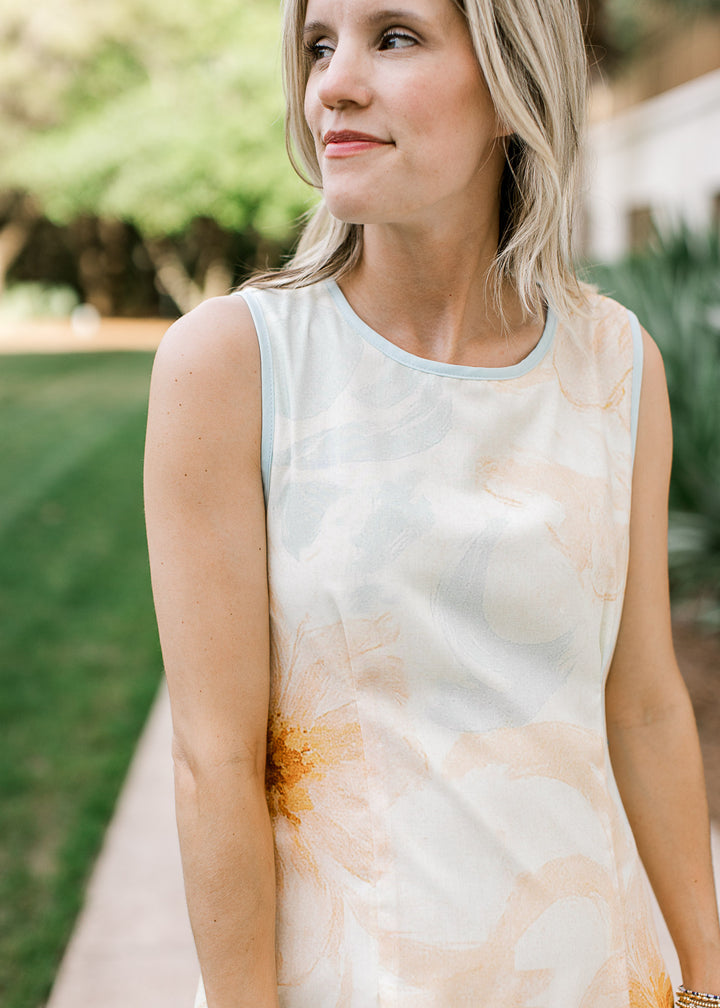 Model wearing a sleeveless cream dress with orange and baby blue floral and baby blue trim. 