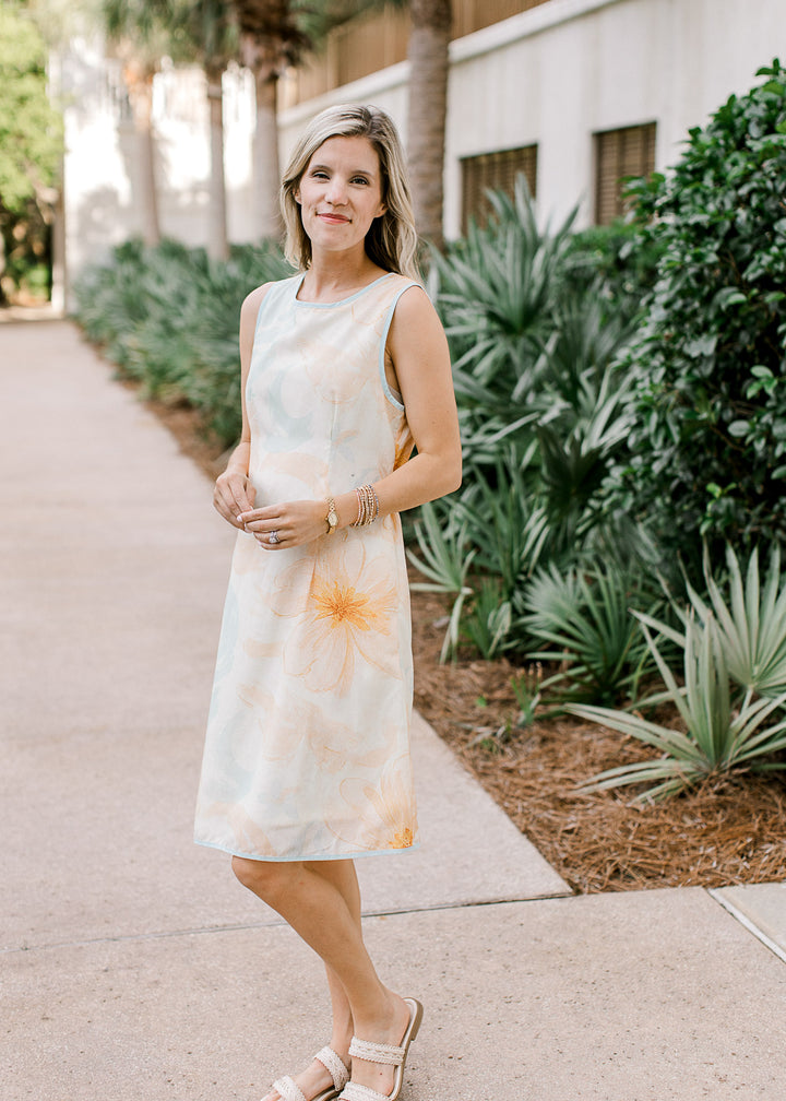 Model wearing sandals and a sleeveless cream dress with orange and baby blue floral and blue trim. 