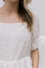 Model wearing a white top adorned white and yellow daises, a square neckline and short poet sleeves.