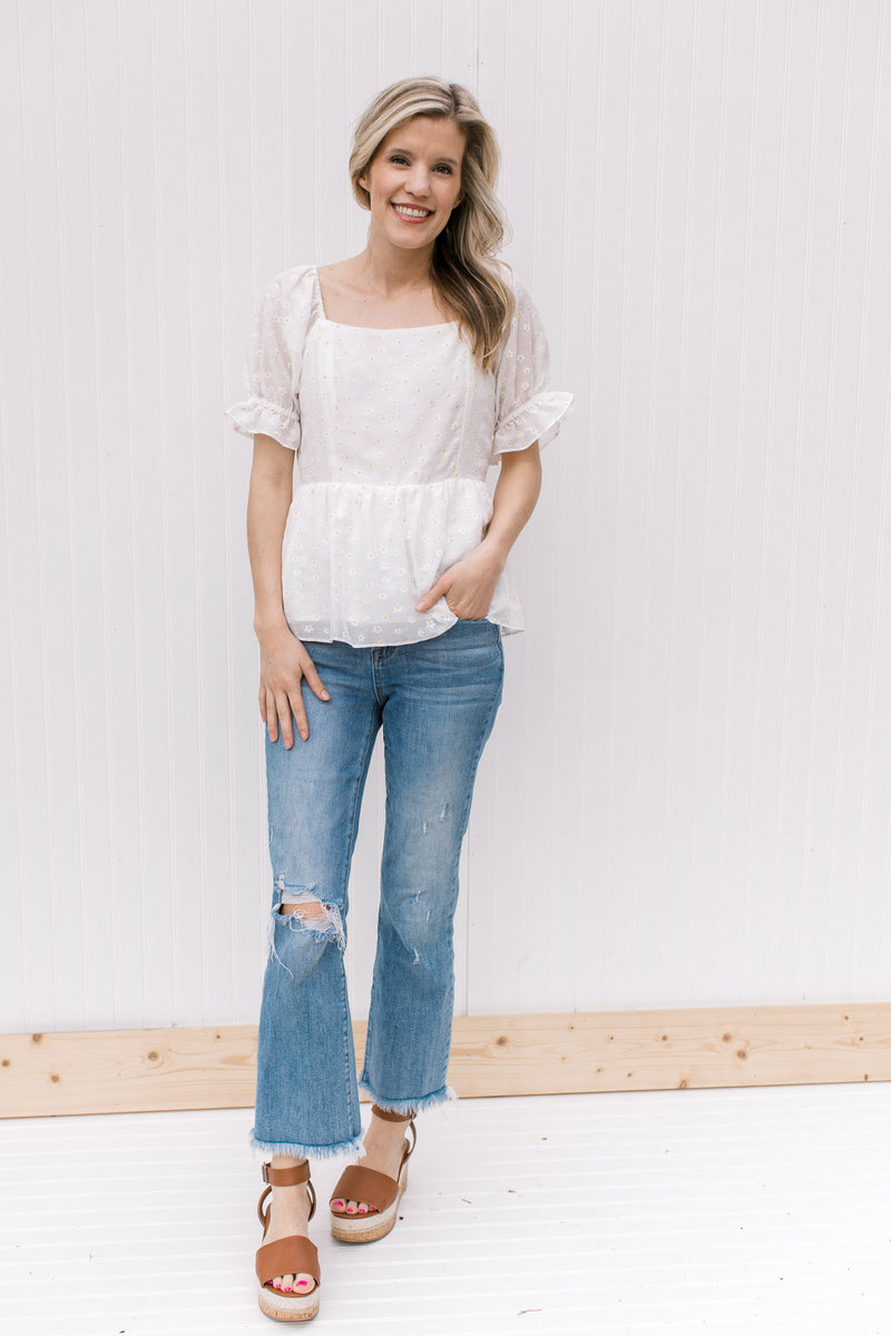 Model wearing jeans, wedges and a white top with a square neck and white and yellow daisies. 