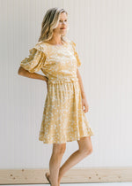 Model wearing a mustard colored above the knee dress with a cream floral pattern and short sleeves.