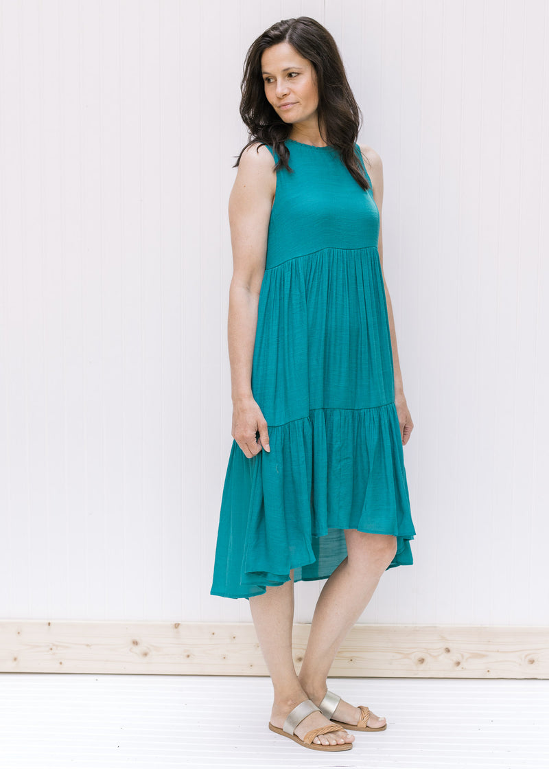Model wearing a deep teal tiered sleeveless dress with a high low silhouette. 