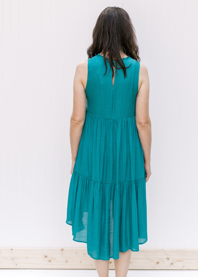 Back view of a Model wearing a deep teal tiered sleeveless dress with a high low silhouette. 