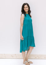 Model wearing sandals with a deep teal tiered sleeveless dress with a high low silhouette. 