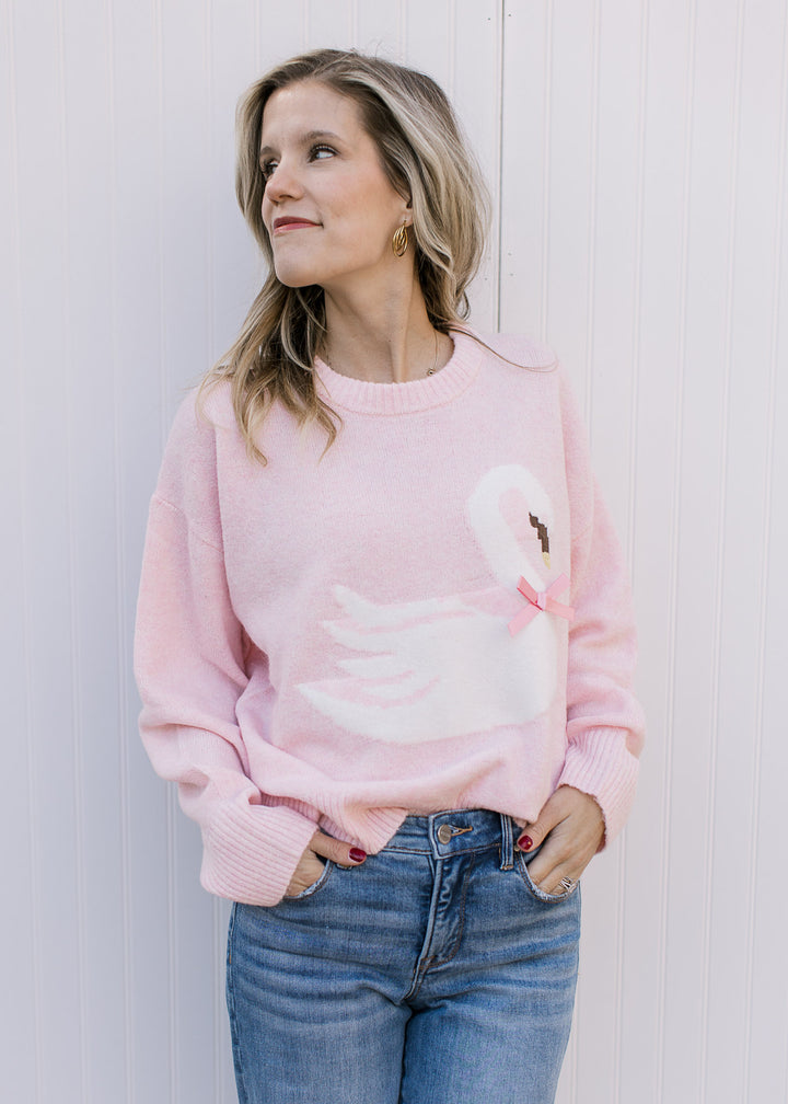 Model wearing a long sleeve pink sweater with a white swan, stitched beak and pink bow detail.