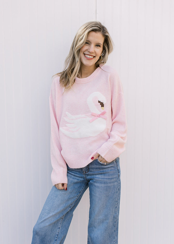 Model wearing jeans and a long sleeve pink sweater with a white swan, stitched beak and pink bow.