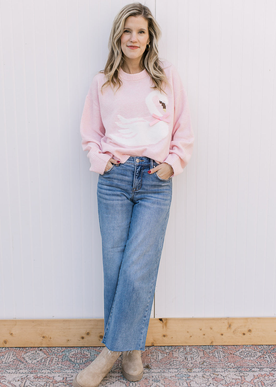 Model wearing jeans, boots and a long sleeve pink sweater with a white swan across the front. 