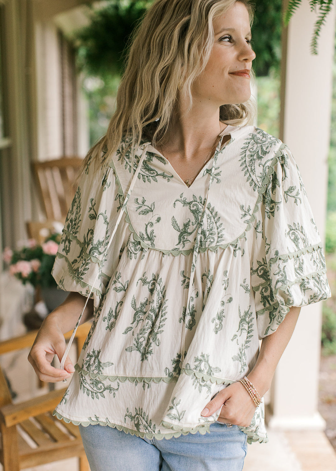 Model wearing a cream top with olive floral pattern, sage scalloped trim and a v-neck with a tie.