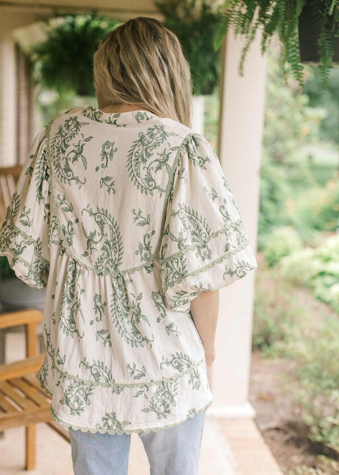 Back view of Model wearing a cream top with olive floral pattern, sage scalloped trim and a v-neck.