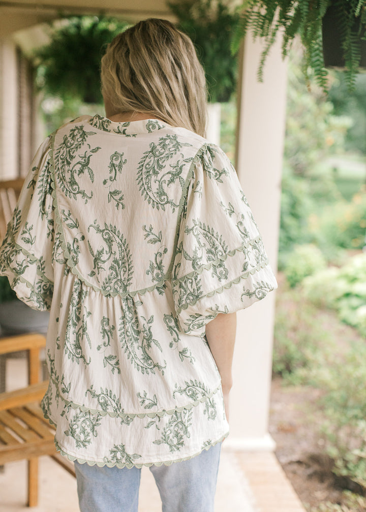 Back view of Model wearing a cream top with olive floral pattern, sage scalloped trim and a v-neck.