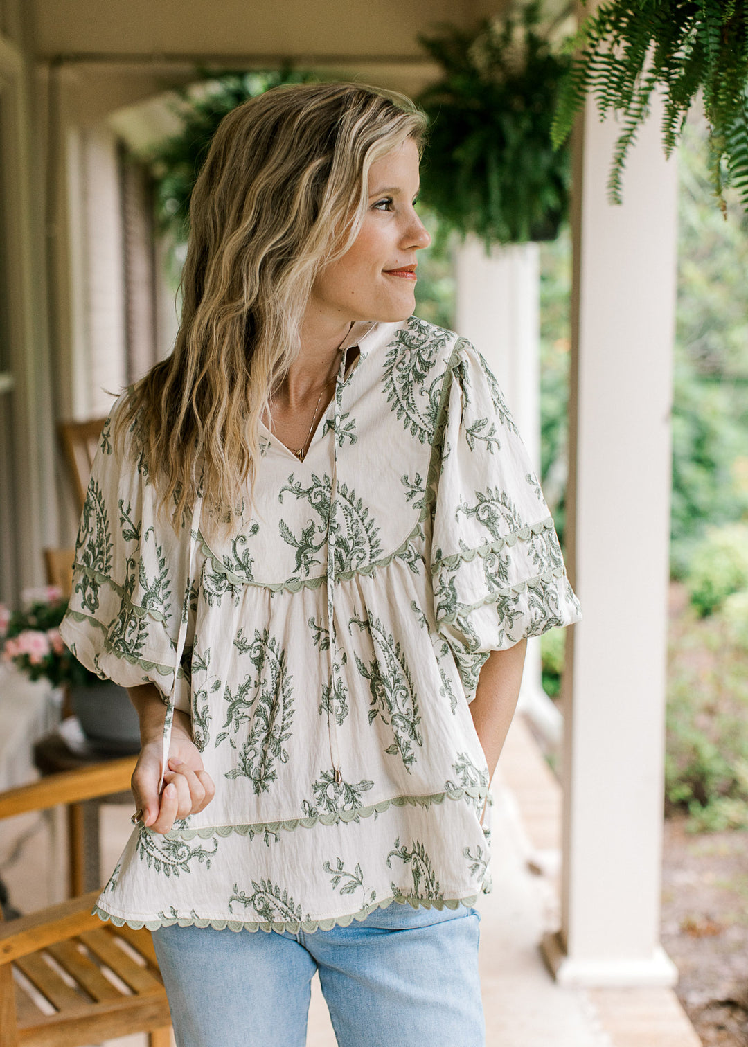 Model wearing jeans and a cream top with olive floral pattern, sage scalloped trim and a v-neck.