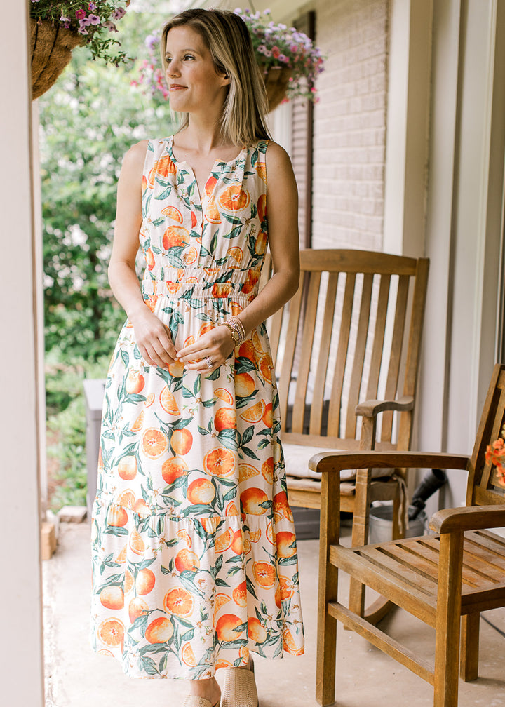 Model wearing a cream sleeveless midi with oranges and green leaf print, v-neck and smocked waist. 