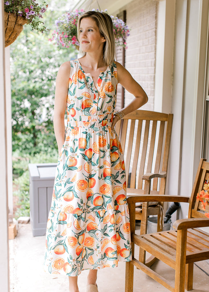 Model wearing wedges and a cream sleeveless midi with oranges and green leaf print. 