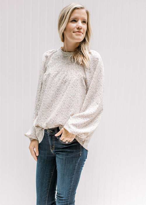 Model wearing jeans with a cream top with a neutral ditsy floral, smocked detail and long sleeves. 