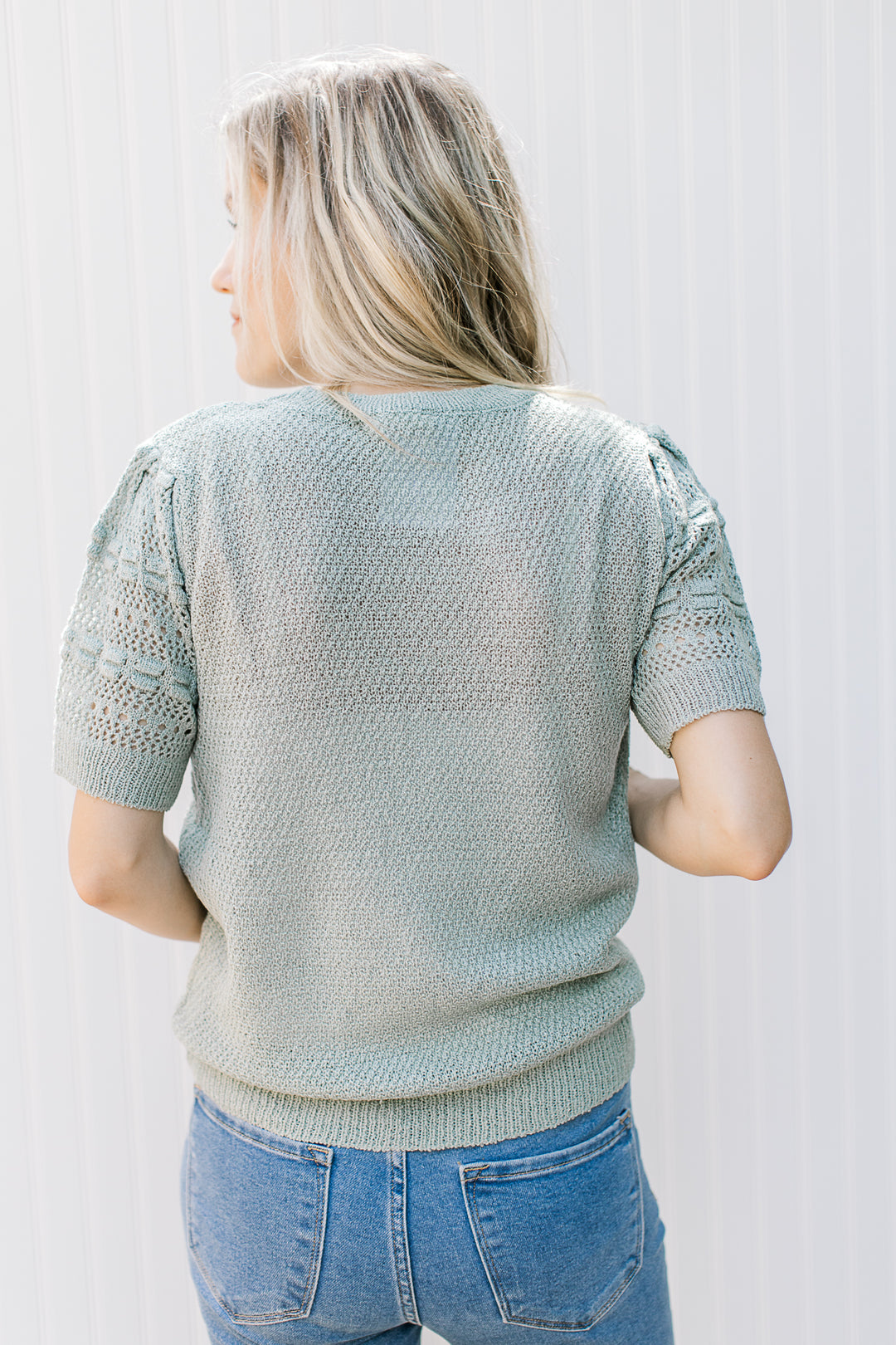 Back view of Model wearing a knit sage sweater with a round neck and eyelet detailed short sleeves. 
