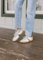 Model wearing cream faux leather body sneakers with suede uppers and gold trim. 