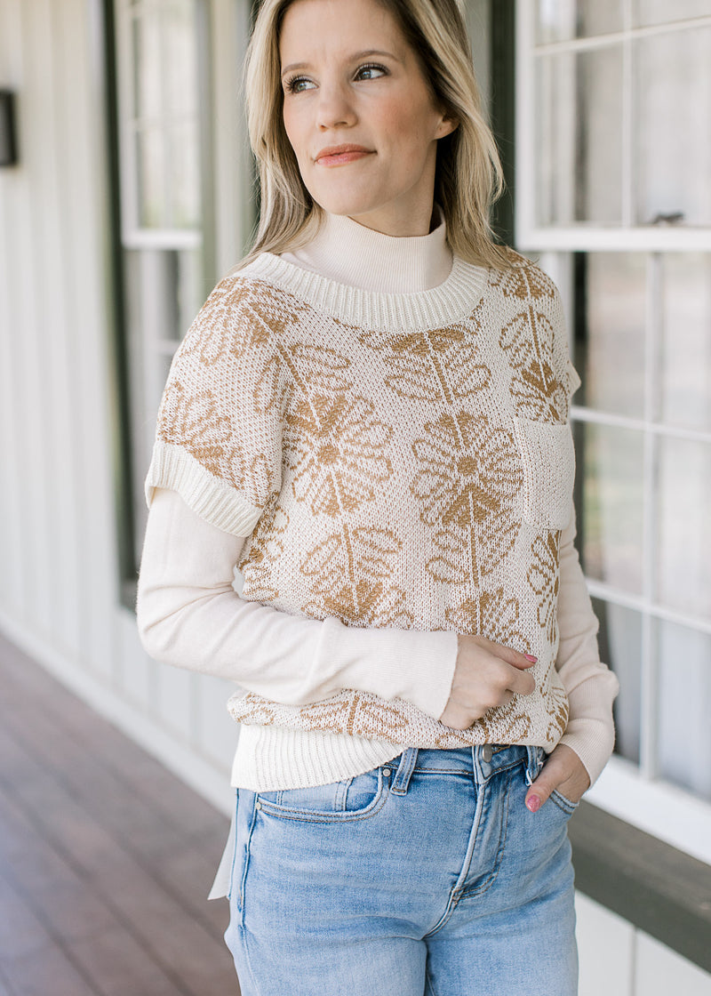 Model wearing a cream mockneck top under a cream sweater with tan flowers and patch pocket.