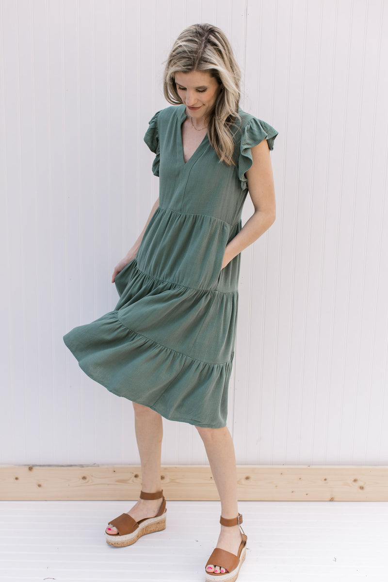 Model wearing wedges with a hunter green tiered midi with ruffle cap sleeves and pockets.