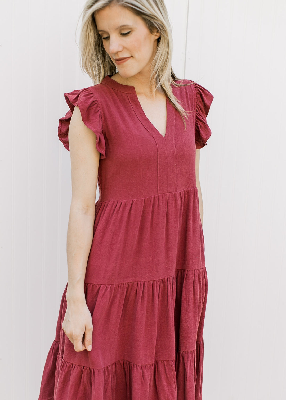 Model wearing a maroon knee length dress with a v-neck, ruffle cap sleeves and a tiered, flowy fit.