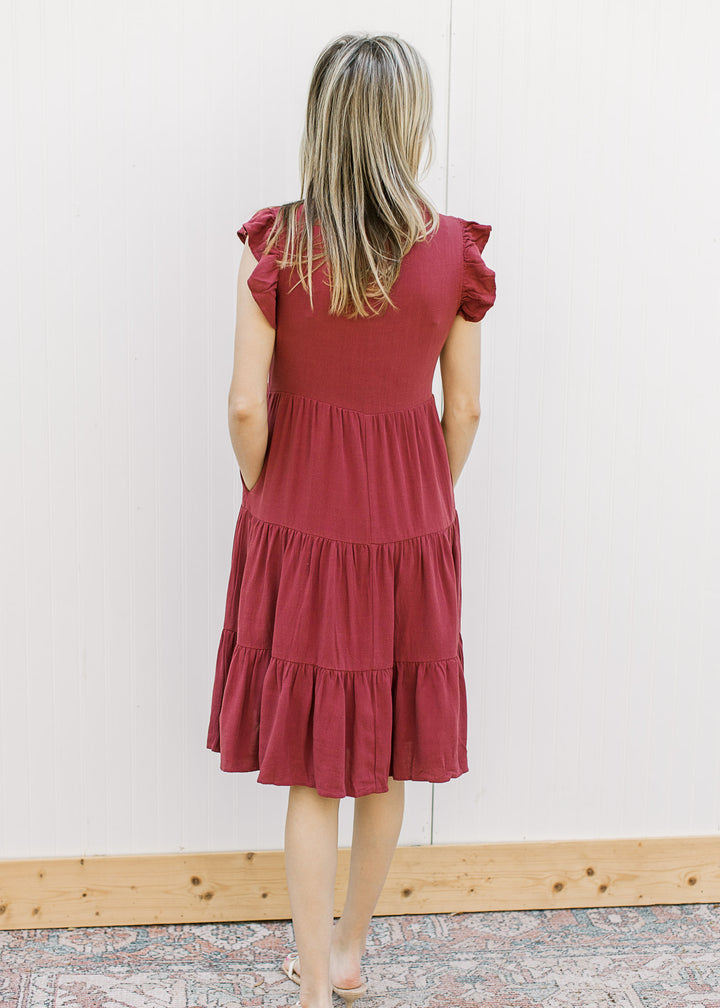 Back view of Model wearing a maroon knee length dress with ruffle cap sleeves and a tiered fit.