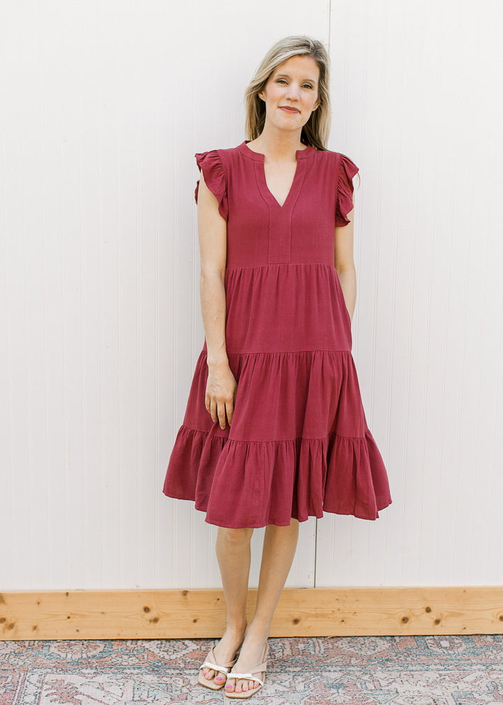 Model wearing heels and a maroon dress with a v-neck, ruffle cap sleeves and a tiered, flowy fit.