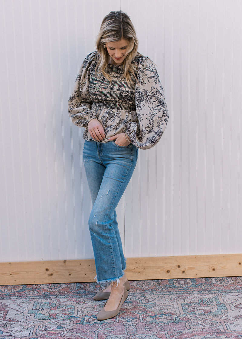 Model wearing jeans and a taupe with black floral top with a smocked bodice and long puff sleeve.