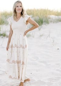 Model is wearing a cream v-neck dress with taupe floral, smocked bodice and ruffle cap sleeve. 