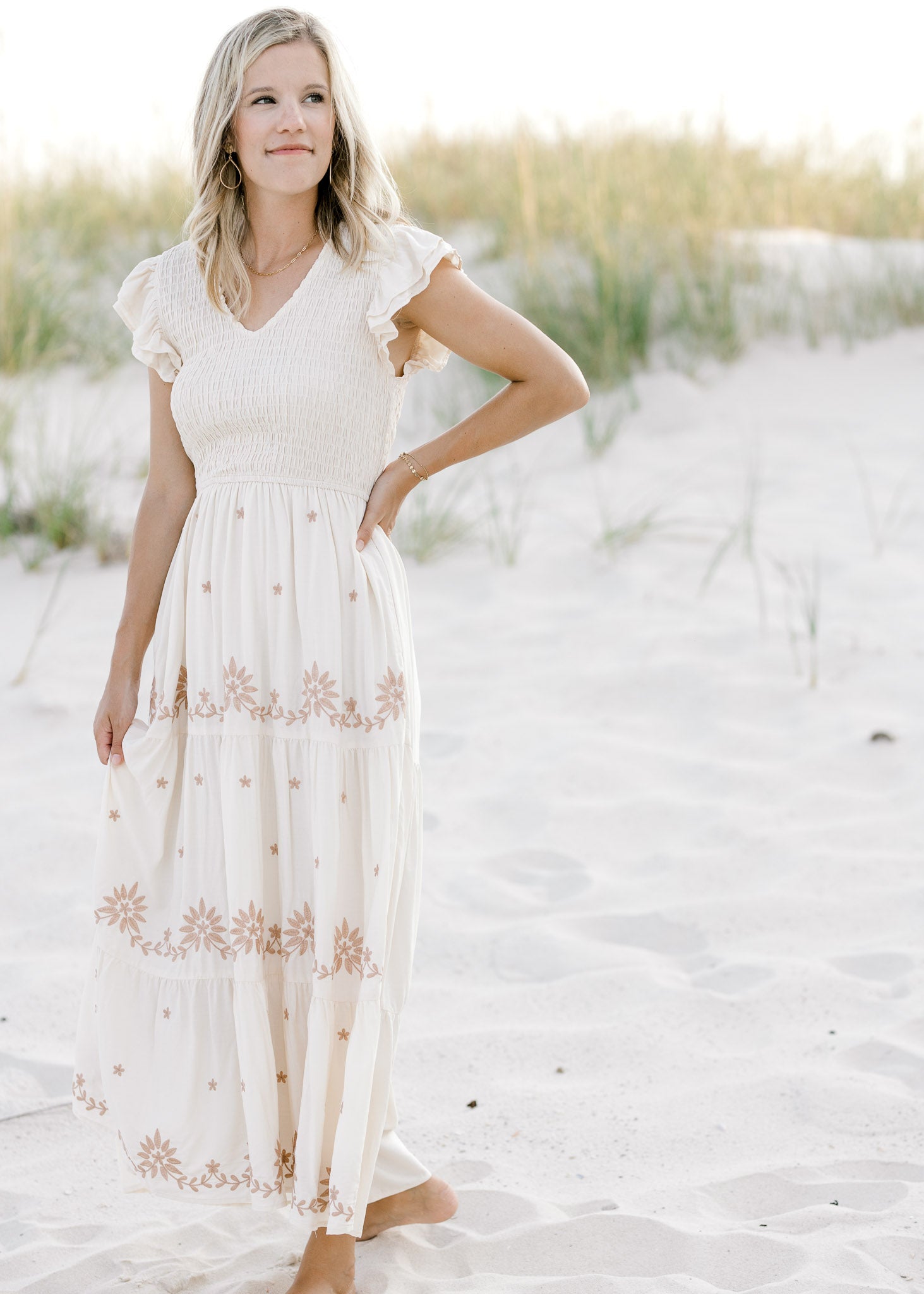 Model is wearing a cream v-neck dress with taupe floral, smocked bodice and ruffle cap sleeve. 