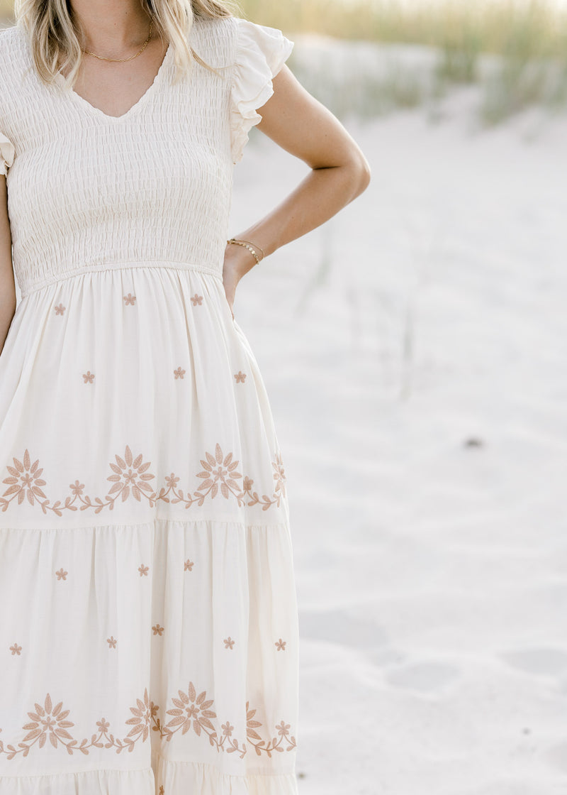 Close up view of smocked bodice and ruffle cap sleeve on a cream dress with taupe embroidered floral