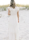 Back view of Model is wearing a cream dress with taupe embroidered floral and ruffle cap sleeve. 