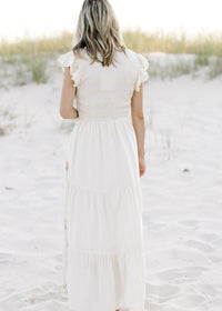 Back view of Model is wearing a cream dress with taupe embroidered floral and ruffle cap sleeve. 