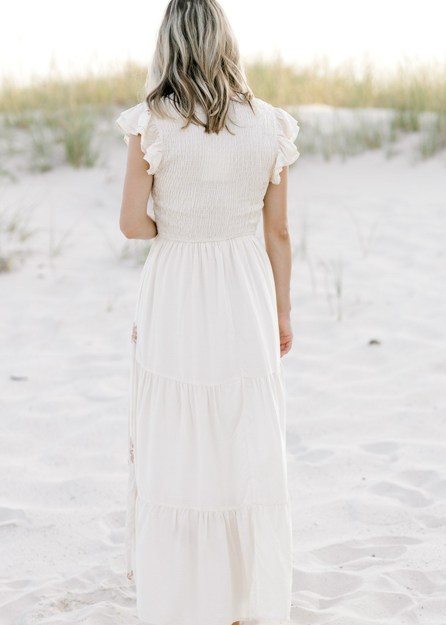 Back view of Model is wearing a cream dress with taupe embroidered floral and ruffle cap sleeve. 