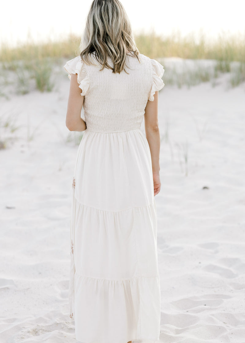 Back view of Model is wearing a cream dress with taupe embroidered floral and ruffle cap sleeve. 