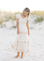 Model is wearing a cream dress with taupe embroidered floral, smocked bodice and ruffle cap sleeve. 