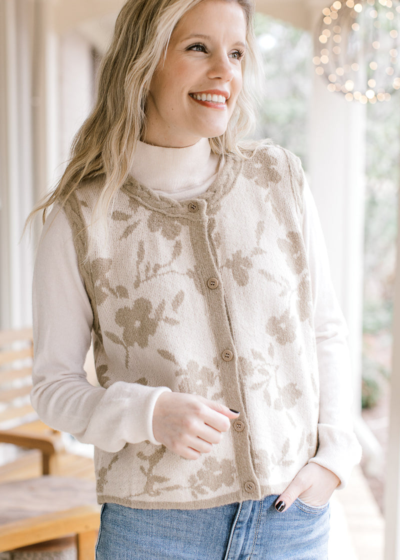 Model wearing a cream sleeveless cardigan with taupe flowers and edging and button front.