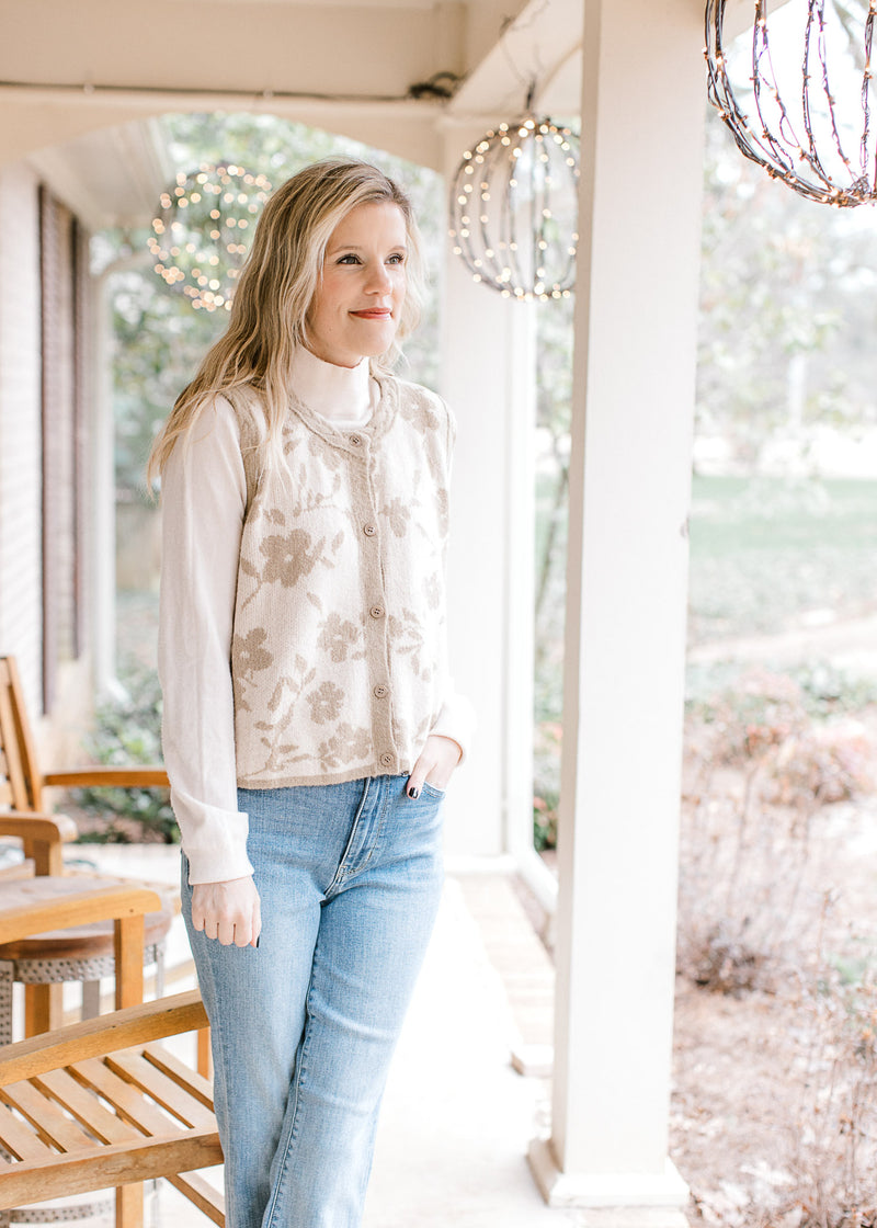 Model wearing jeans and a cream sleeveless cardigan with taupe flowers and edging.