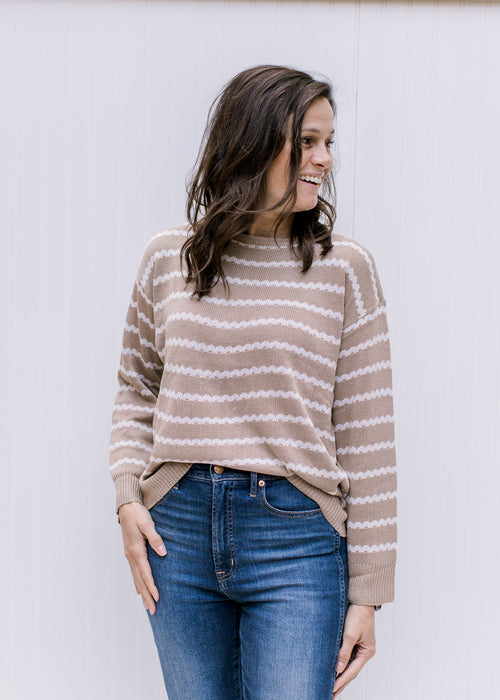 Model wearing a taupe sweater with cream zig zag stripes, a round neck and a zipper in the back. 