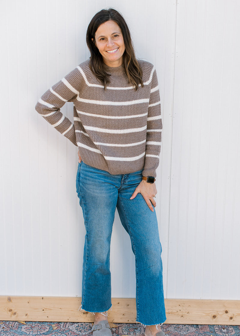 Model wearing jeans, clogs and a taupe sweater with white stripes and long sleeves.