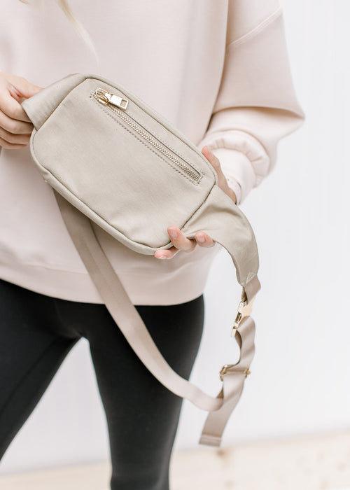 Model holding a gold and taupe belt bag with an adjustable strap and three zipper pockets. 
