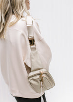 Model with a gold and taupe belt bag with an adjustable strap and zipper pockets over her shoulder. 
