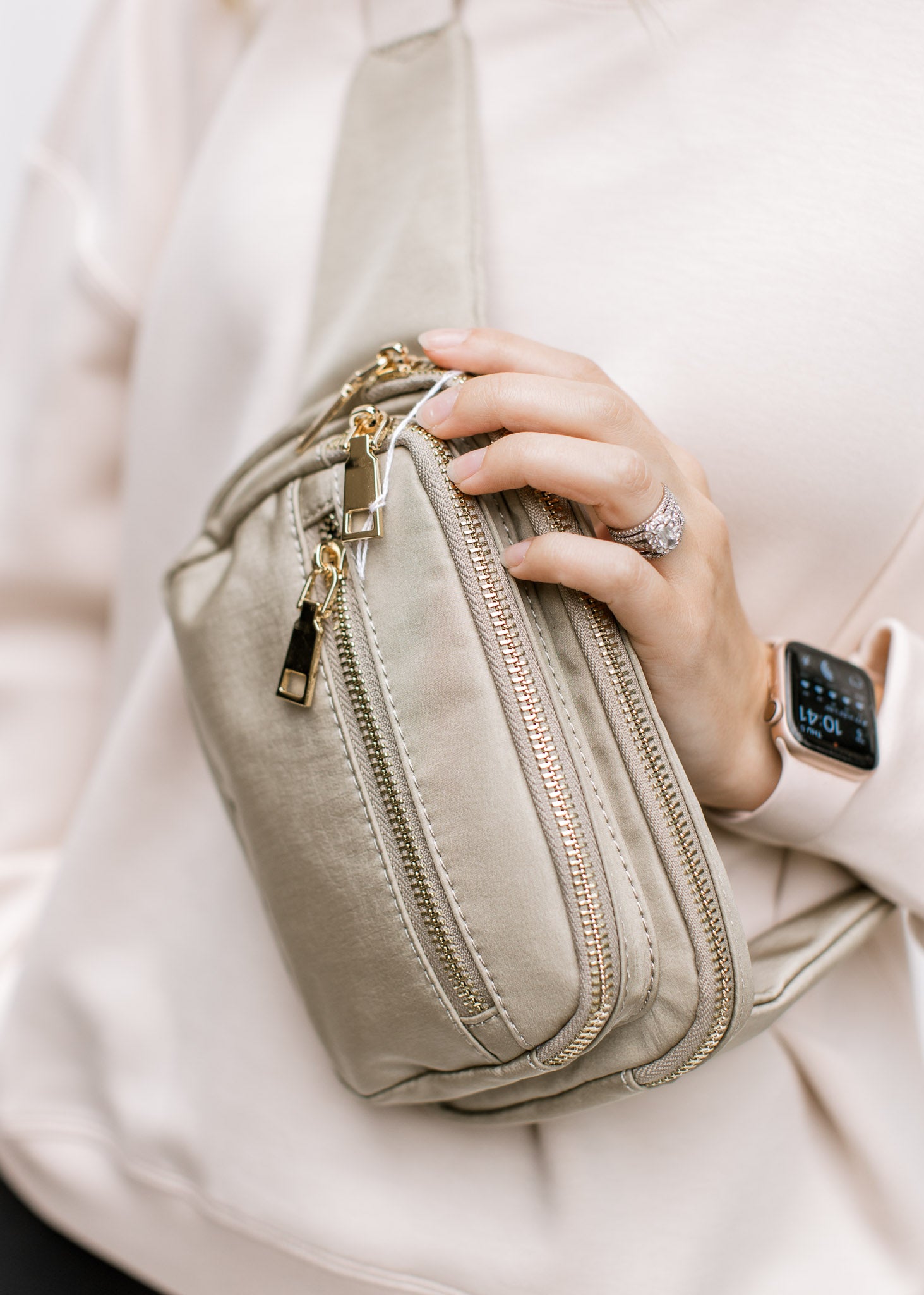 Close up of Model holding a gold and taupe belt bag with an adjustable strap and zipper pockets. 