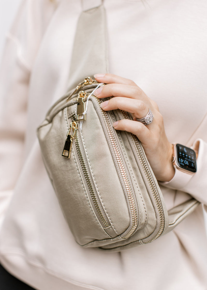 Close up of Model holding a gold and taupe belt bag with an adjustable strap and zipper pockets. 